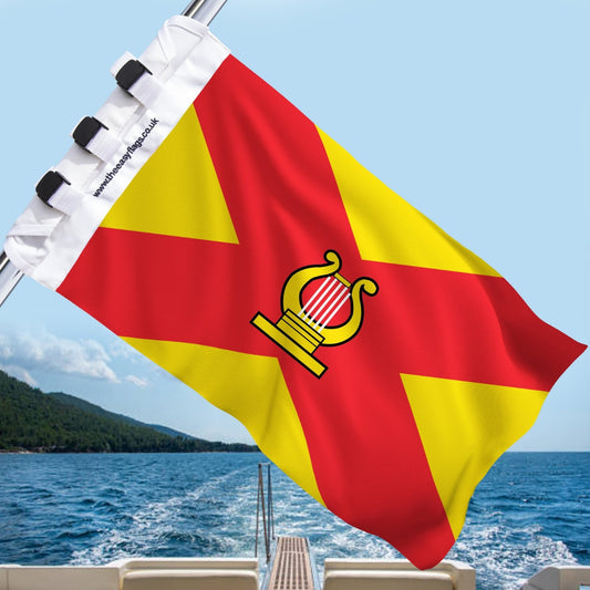 Ayrshire county flag with red background and bold yellow saltire, featuring a golden harp emblem at the centre, mounted on a boat rail with a universal triple-lock system, flying at sea with blue sky and coastline in the background