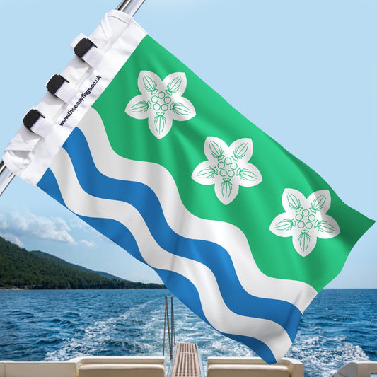 Cumberland county flag with green background, three white parnassus flowers, and blue and white wavy stripes, mounted on a boat rail with a universal triple-lock system, flying at sea with blue sky and coastline in the background