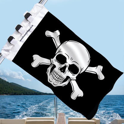 Black Jolly Roger pirate flag with skull and crossbones, mounted on a boat rail with a universal triple-lock system, flying at sea with blue sky and coastline in the background