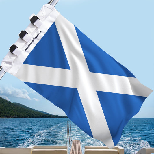Scotland Saltire flag with white diagonal cross on blue background, mounted on a boat rail with a universal triple-lock system, flying at sea with blue sky and coastline in the background