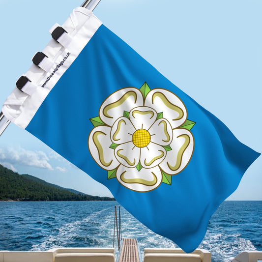 Yorkshire county flag with blue background and white rose emblem outlined in gold, with green sepals and yellow centre, mounted on a boat rail with a universal triple-lock system, flying at sea with blue sky and coastline in the background